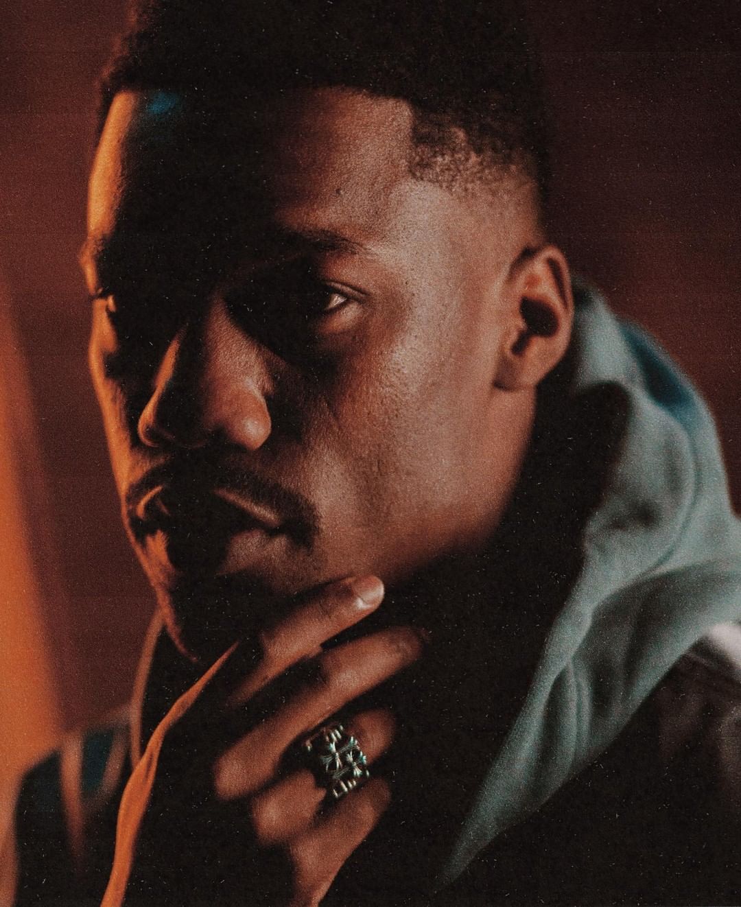Close-up portrait of an African American man with a hand near his face, wearing a hooded jacket and a distinctive ring, in warm-toned lighting.