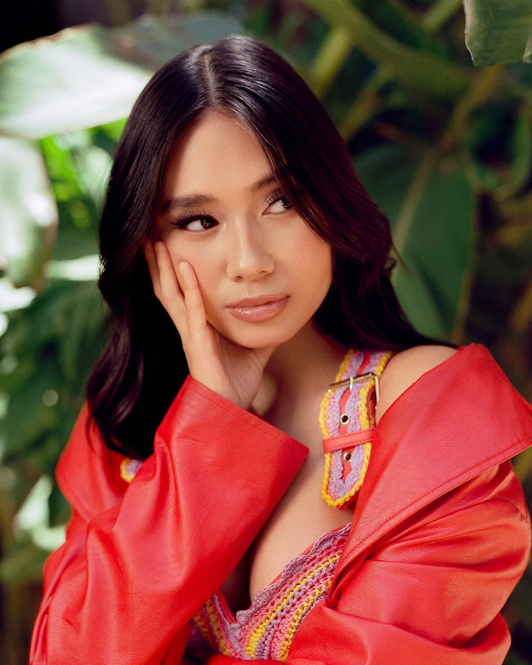 Portrait of an Asian woman in a red jacket with dark hair, posing with her hand near her face against a foliage background.