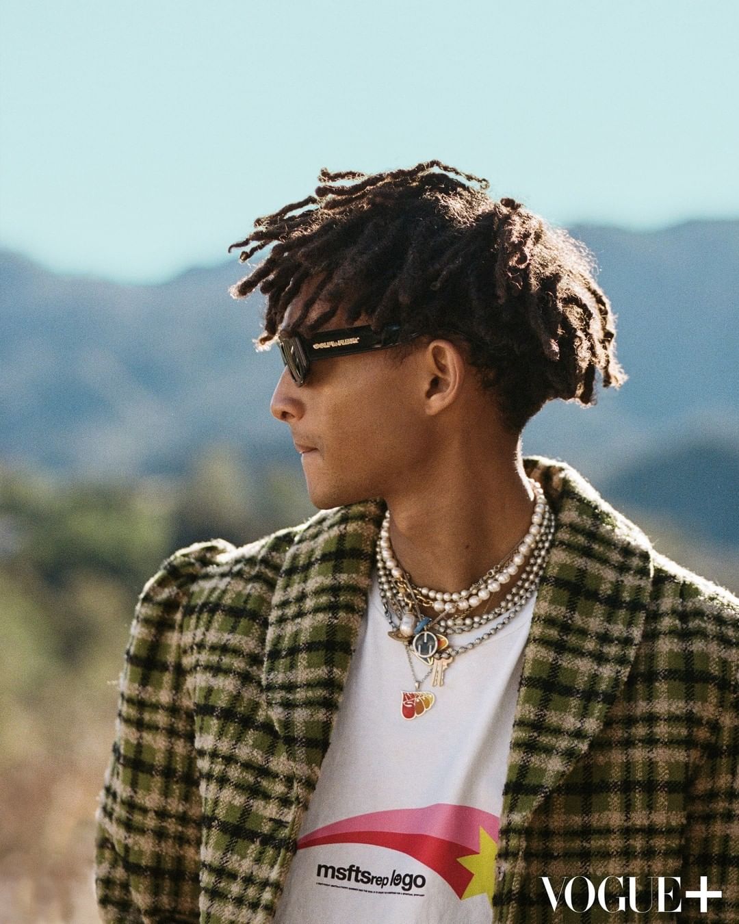 Man with dreadlocks, sunglasses, plaid jacket, and layered pearl necklaces in an outdoor portrait against a mountain backdrop.