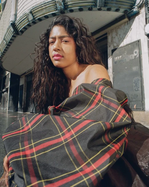 Young woman with long dark hair wearing a red and black plaid top sitting in front of a concrete building on a cloudy day.