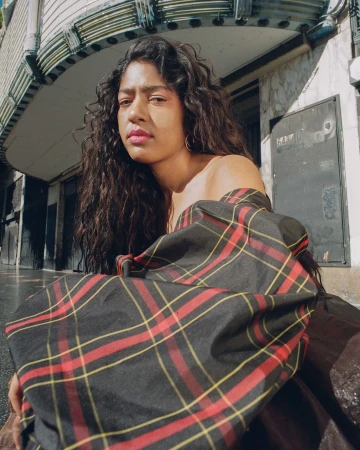 Young woman with long dark hair wearing a red and black plaid top sitting in front of a concrete building on a cloudy day.