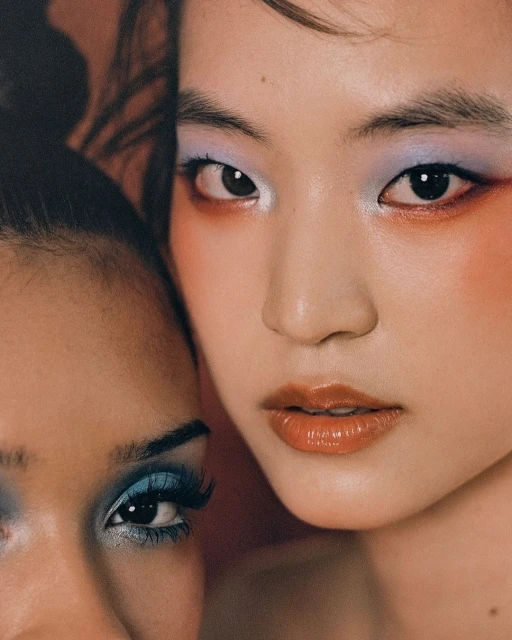 Close-up portrait of two diverse young adult females with colorful make-up in peachy studio lighting showcasing cosmetic beauty.