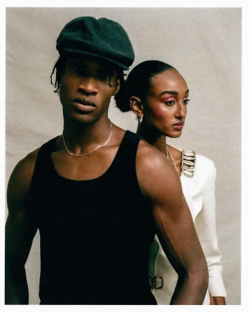 Portrait of a Black man in a black tank top and flat cap with a woman in a white jacket standing behind him against a neutral textured wall