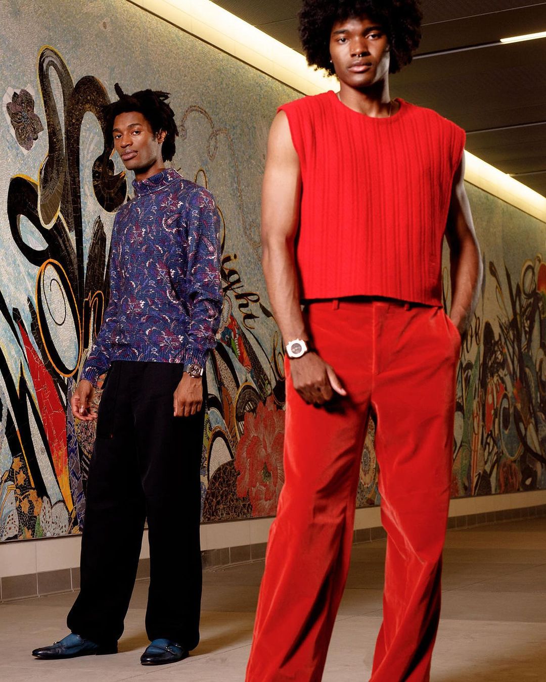 Two black men posing in a mosaic-tiled corridor photographed by Alex Harper, one in a red sleeveless top and red trousers.