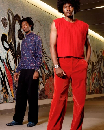 Two black men posing in a mosaic-tiled corridor photographed by Alex Harper, one in a red sleeveless top and red trousers.
