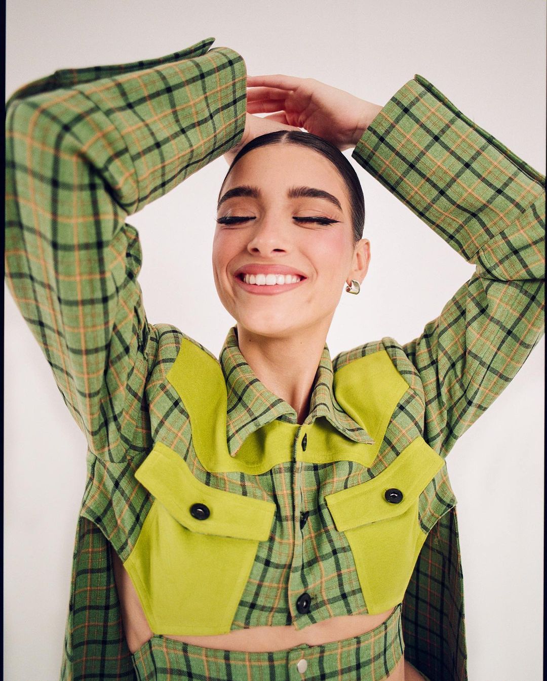Smiling woman in a green plaid jacket posing with arms raised, photographed by Alex Harper against a plain background