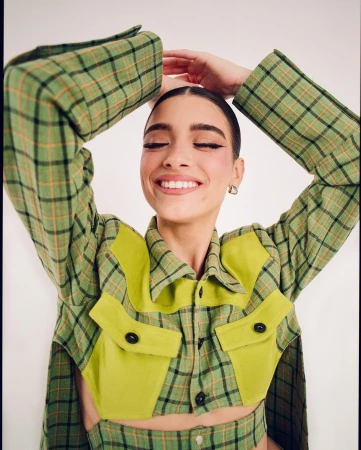 Smiling woman in a green plaid jacket posing with arms raised, photographed by Alex Harper against a plain background