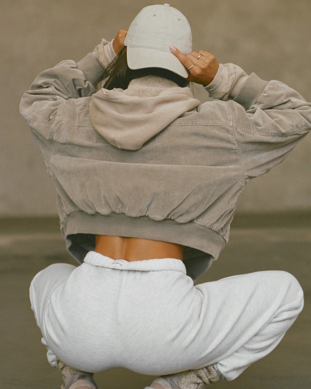 Woman in casual wear adjusts cap, featuring a hoodie, jacket, and sweatpants. Low angle in neutral color tones. Fashion shoot.