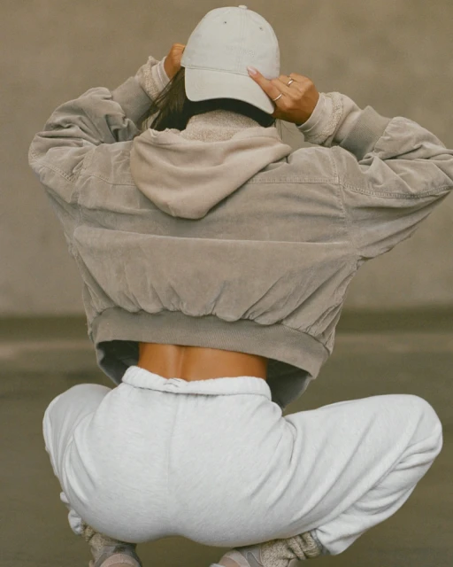 Woman in casual wear adjusts cap, featuring a hoodie, jacket, and sweatpants. Low angle in neutral color tones. Fashion shoot.