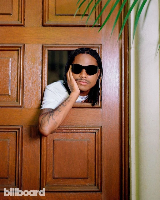Man with dreadlocks, sunglasses posing in a doorway with hand on cheek; indoor shot with houseplant and 'billboard' logo. 