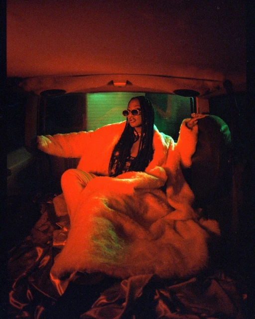 Portrait of an adult female with braids and sunglasses, reclining in a car trunk amidst faux fur and draped satin fabric, bathed in red lighting.