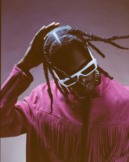 Portrait of Black adult male with braids and sunglasses in a magenta fringe jacket against a muted purple background.