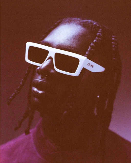 Close-up studio portrait of an African-American adult male wearing white-framed sunglasses with dark lenses against a brown backdrop.