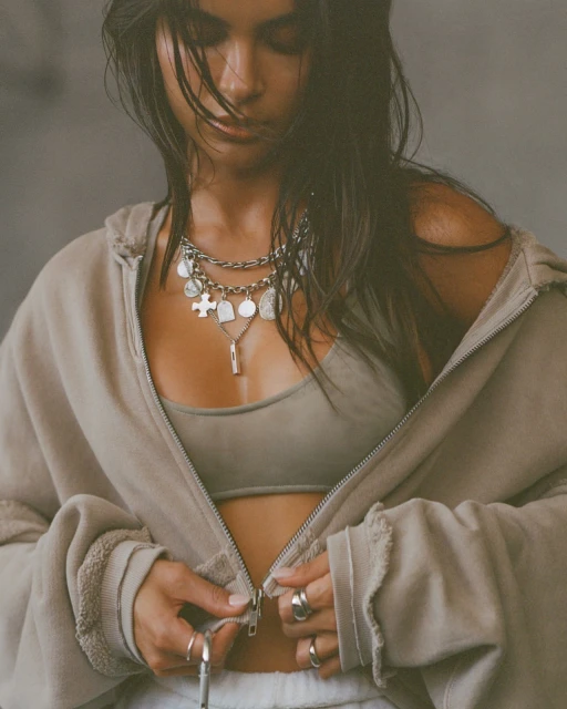 Woman wearing a silver charm necklace and beige hoodie is shown in a medium shot, slightly angled down with a neutral background.