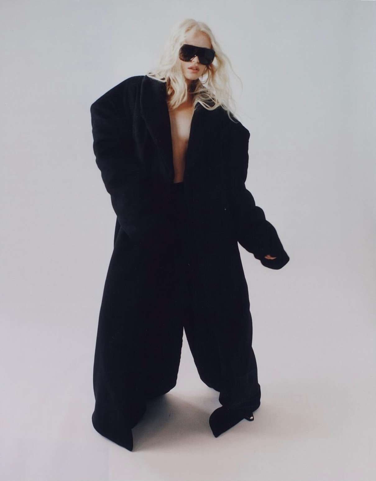 Blonde woman in oversized black coat and sunglasses poses against a white backdrop in a fashion forward and modern style.