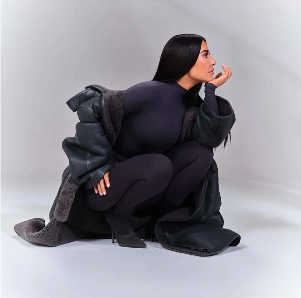 Kim Kardashian squatting in profile wearing a dark turtleneck bodysuit and oversized shearling coat against a white background