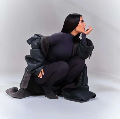 Kim Kardashian squatting in profile wearing a dark turtleneck bodysuit and oversized shearling coat against a white background