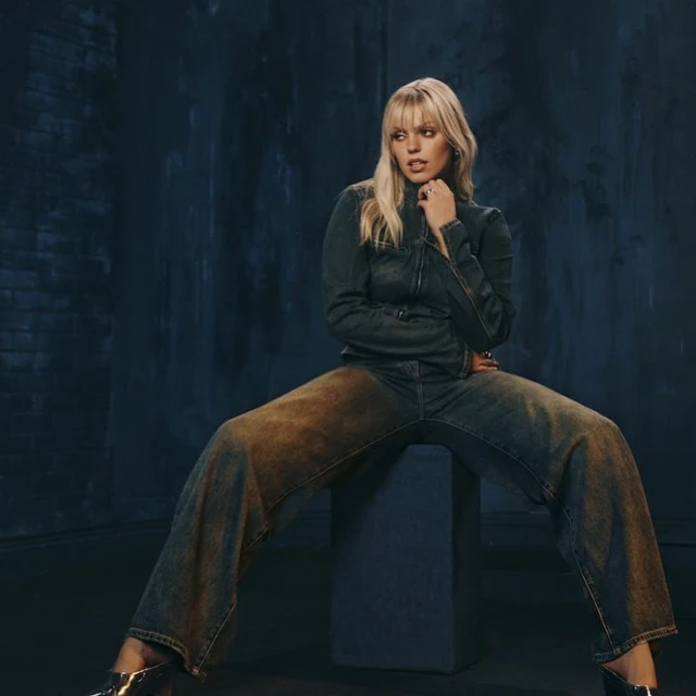 An adult female model sits on a block in a photo studio, wearing a denim jacket and wide-leg jeans with brown distressed details.