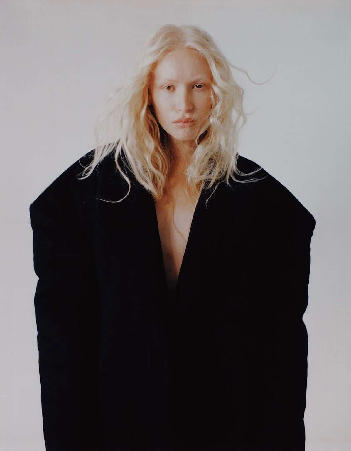 Portrait of a blonde woman wearing an oversized black coat against a plain off-white studio background by photographer Alex Harper.