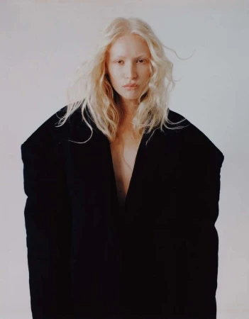 Portrait of a blonde woman wearing an oversized black coat against a plain off-white studio background by photographer Alex Harper.