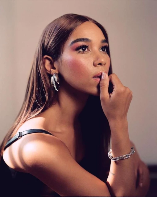 Portrait of a young woman with long brown hair and pink makeup, wearing silver jewelry, looking upward in a softly lit, neutral setting.