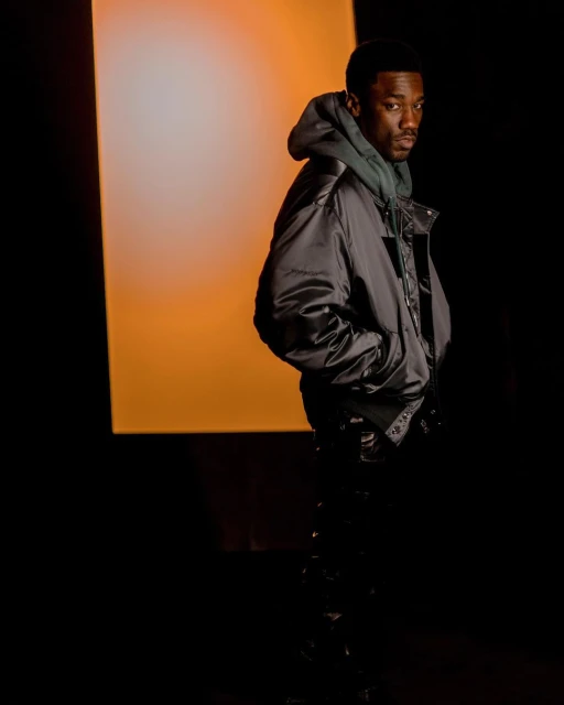 Man in shiny black jacket with hood, hands in pockets, poses against a black backdrop with orange light panel.