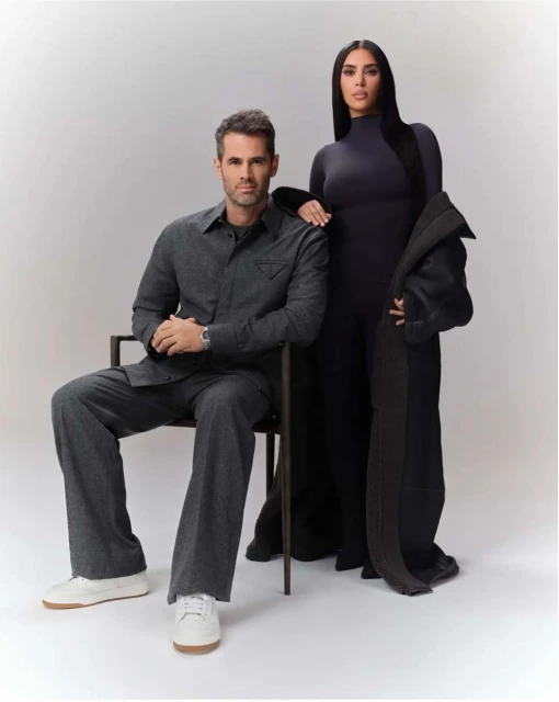 Man seated next to woman in studio; both wearing matching charcoal outfits and neutral sneakers against a gradient backdrop.