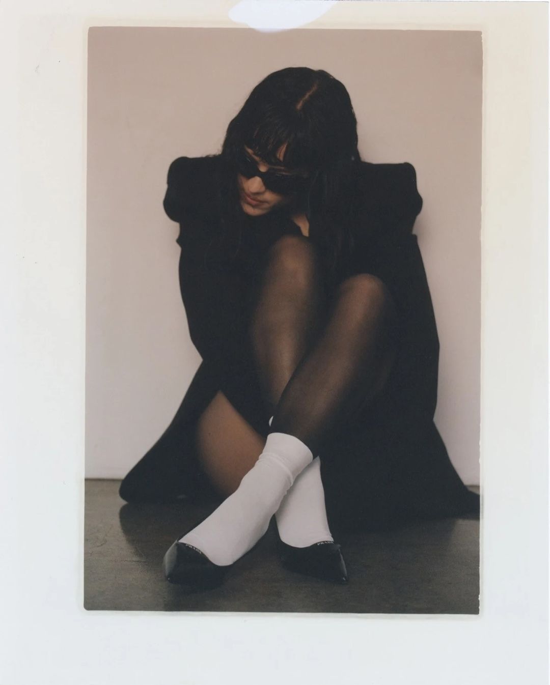 A young woman with dark hair poses indoors wearing a black dress, black stockings, white socks and black flats, sitting against a neutral wall.