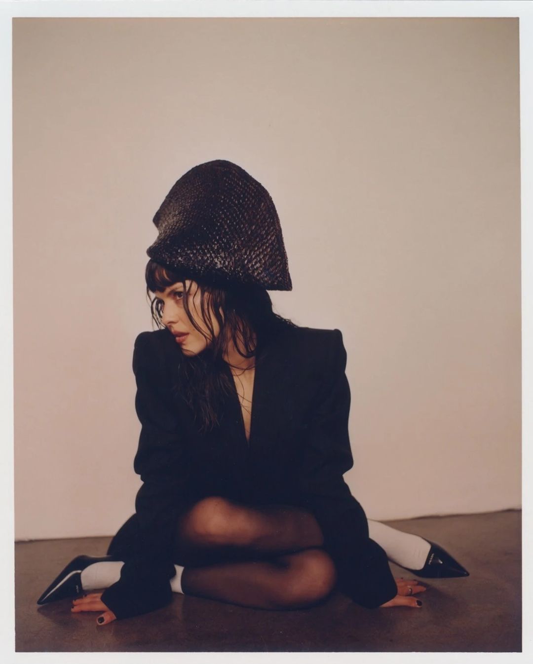 Fashionable woman wearing a black blazer and textured hat poses on the floor in an indoor studio setting with neutral lighting.