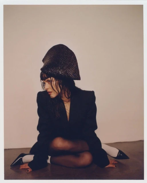 Fashionable woman wearing a black blazer and textured hat poses on the floor in an indoor studio setting with neutral lighting.