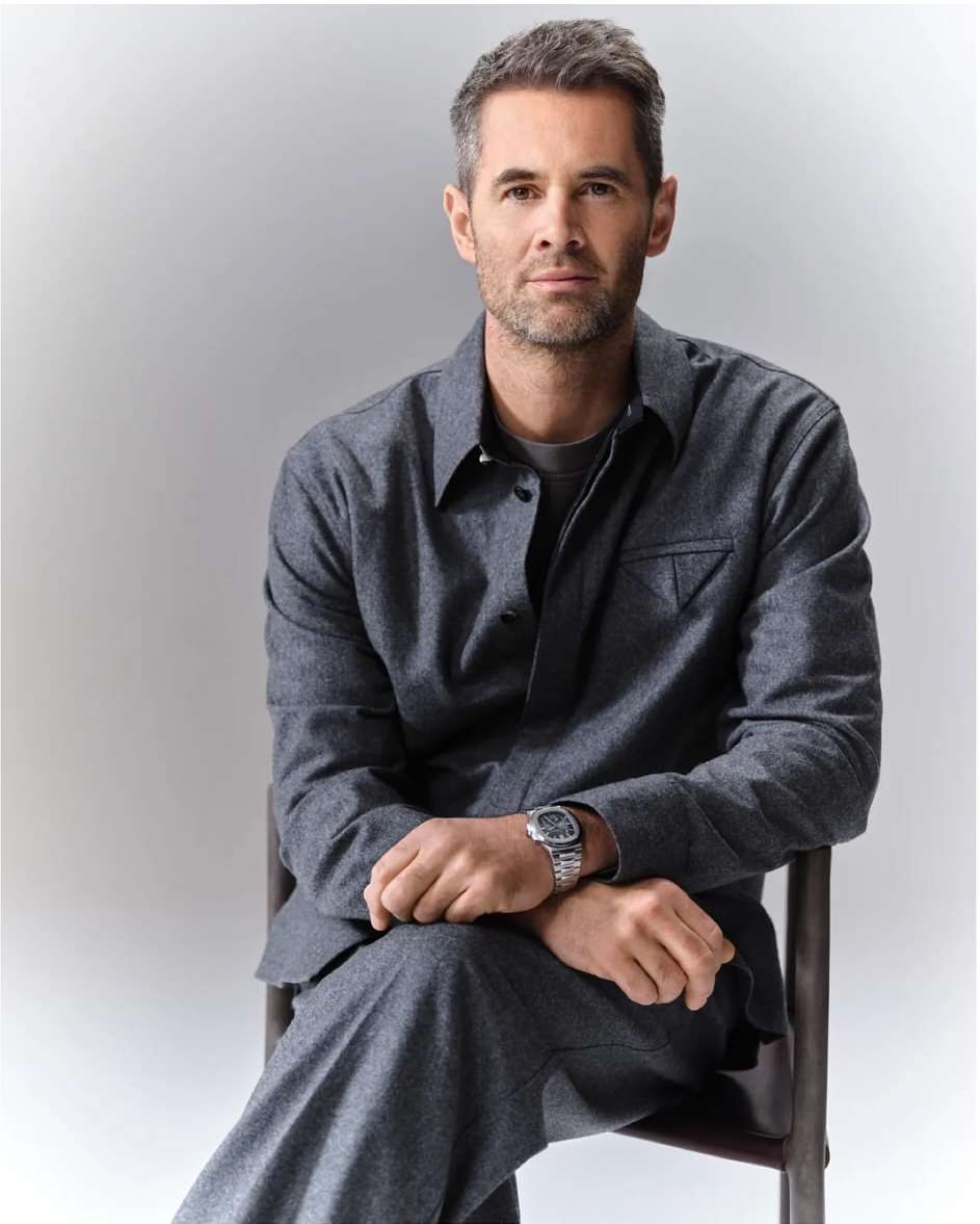 Portrait of an adult male with gray hair and a beard, wearing a gray suit and silver watch, sitting against a light gray backdrop.