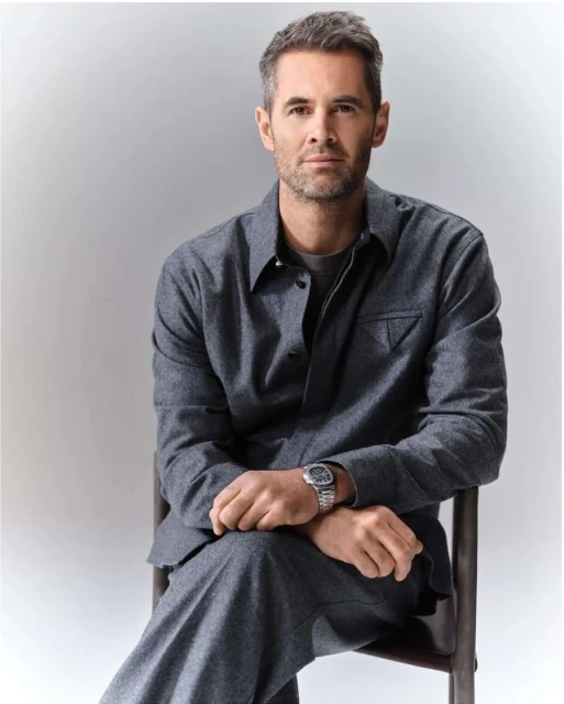 Portrait of an adult male with gray hair and a beard, wearing a gray suit and silver watch, sitting against a light gray backdrop.