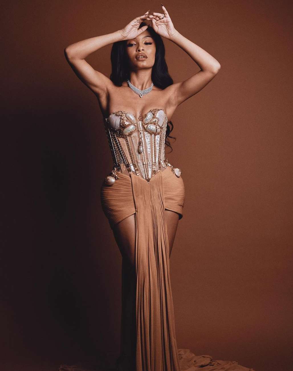 Elegant Black woman in a nude gown with her arms raised posing against a brown backdrop for a high fashion portrait indoors.