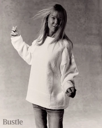Portrait of a blonde woman wearing a textured white sweater and denim jeans against a textured grey background.