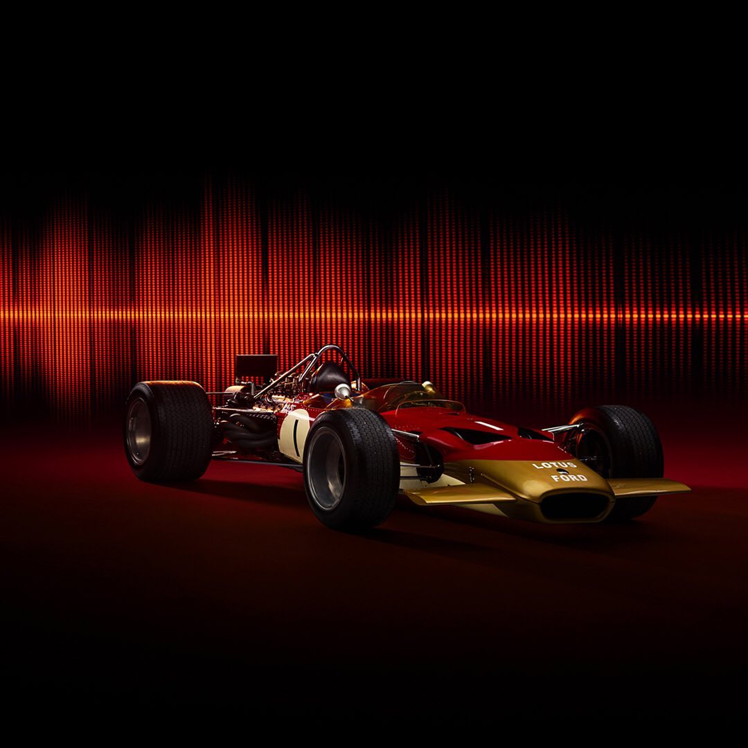 A red and gold vintage Lotus Ford race car, shot in a photo studio with a graphic red background.