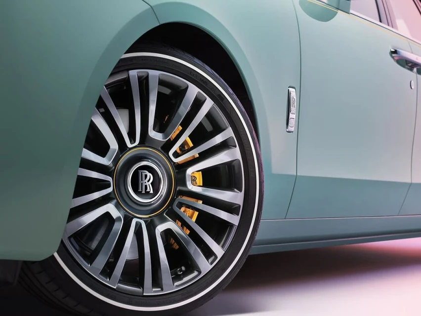 Close-up of a teal Rolls-Royce car featuring a multi-spoke wheel with a prominent 'R' logo and yellow brake calipers against a light background.