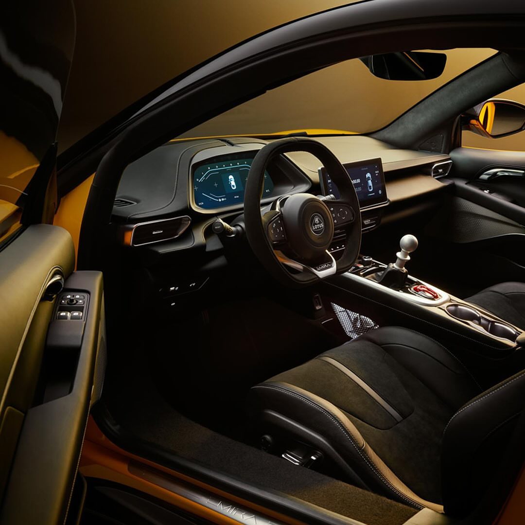 Interior view of an orange Lotus Emira sports car featuring a dashboard display, leather seats, steering wheel, and console details.