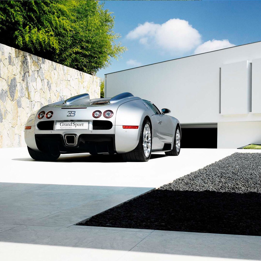 Silver Bugatti Grand Sport parked outside a modern white building with a stone wall and green trees under a blue sky.