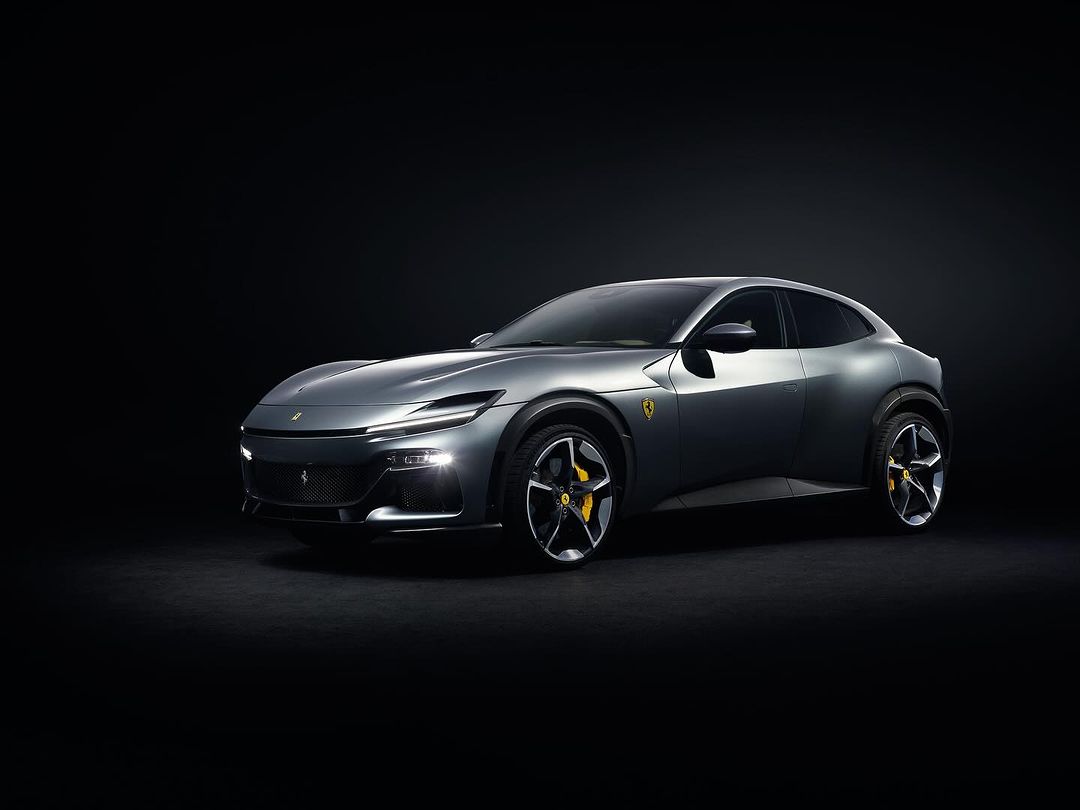 Sleek grey Ferrari Purosangue SUV with yellow brake calipers, illuminated headlights, parked against a stark black backdrop in a studio setting.