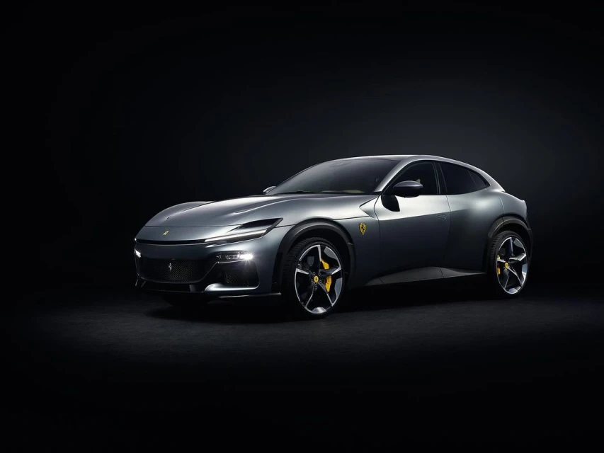 Sleek grey Ferrari Purosangue SUV with yellow brake calipers, illuminated headlights, parked against a stark black backdrop in a studio setting.