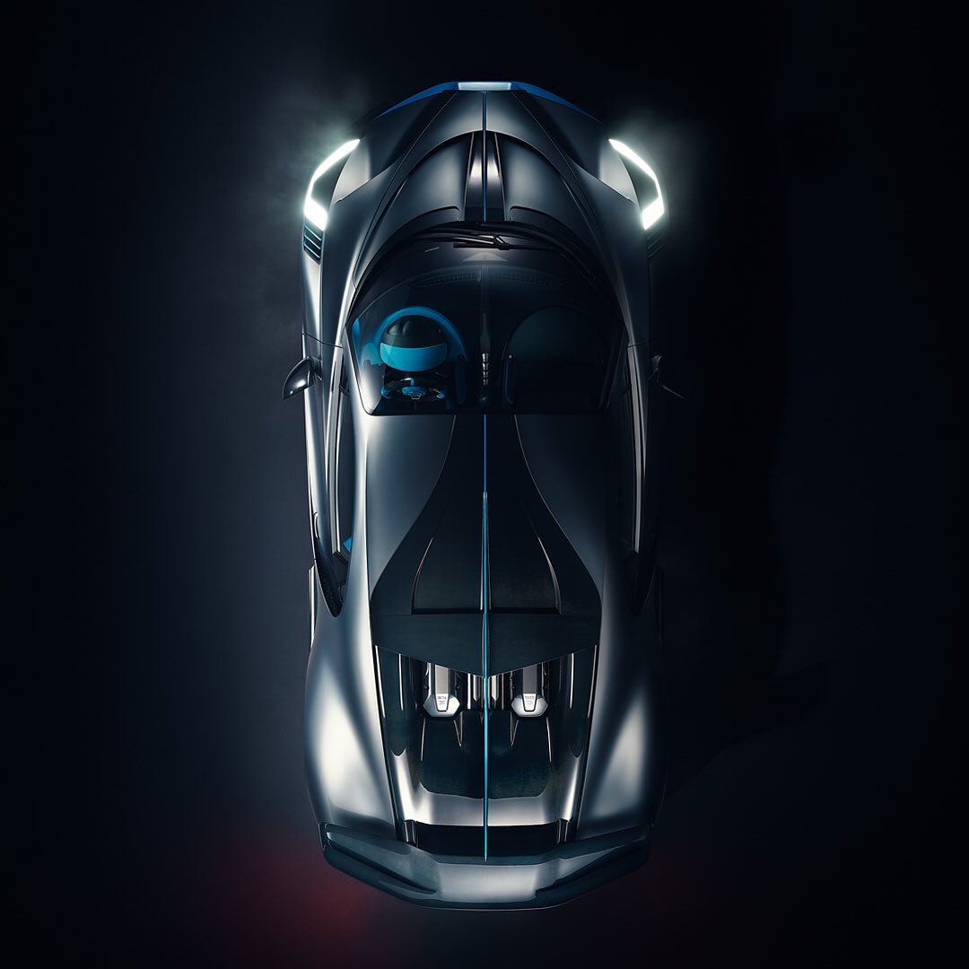 Sleek silver Bugatti sports car seen from an overhead angle on a black background, highlighting its aerodynamic design and illuminated headlights.