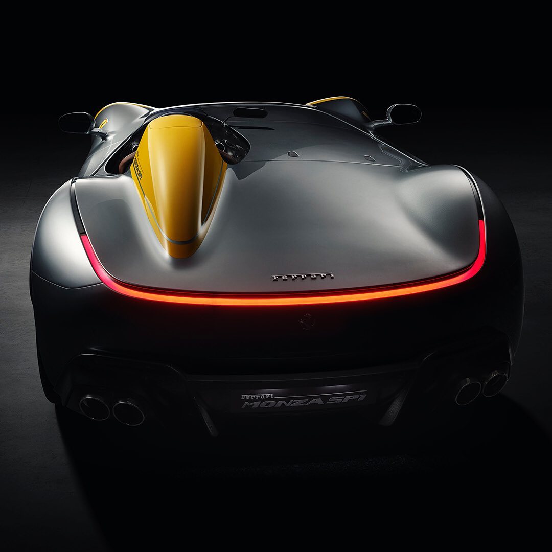 Rear view of a gray and yellow Ferrari Monza SP1 sports car against a black background showcasing its sleek design and taillights.