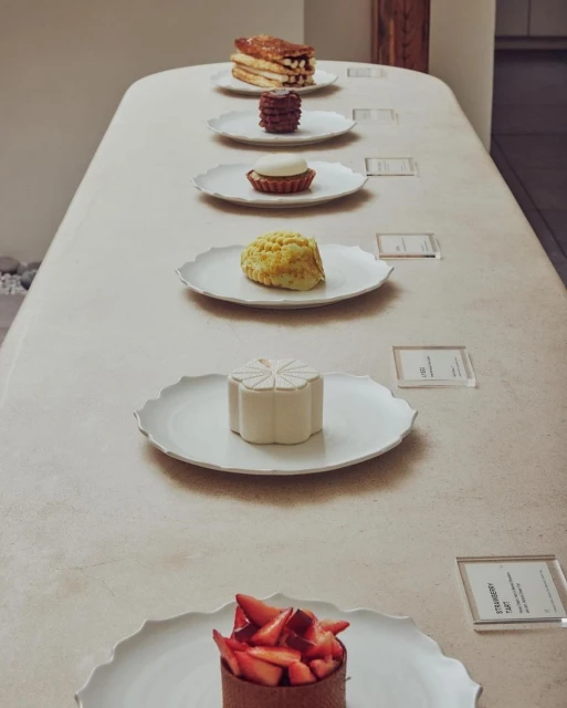 Arrangement of desserts including cakes, tarts, and pastries displayed on a long, light-colored table, each on individual white plates.