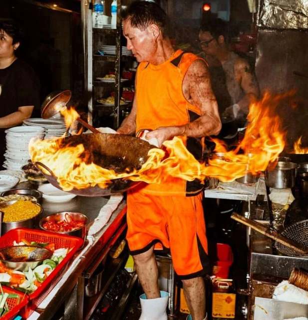 A chef in an orange uniform stir-frying in a wok with flames, cooking in a commercial kitchen with food ingredients and restaurant equipment.