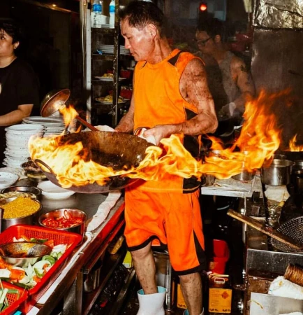 A chef in an orange uniform stir-frying in a wok with flames, cooking in a commercial kitchen with food ingredients and restaurant equipment.