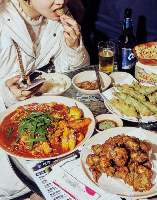 A person eats Korean food at a table with an array of dishes and a beer bottle, set indoors with warm lighting.