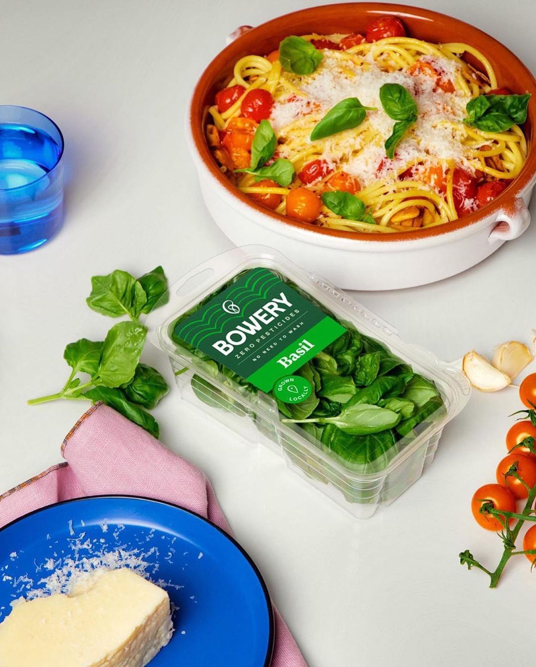 Overhead food photography displays pasta with tomato sauce and basil, container of basil, blue dish, and a glass of blue liquid.