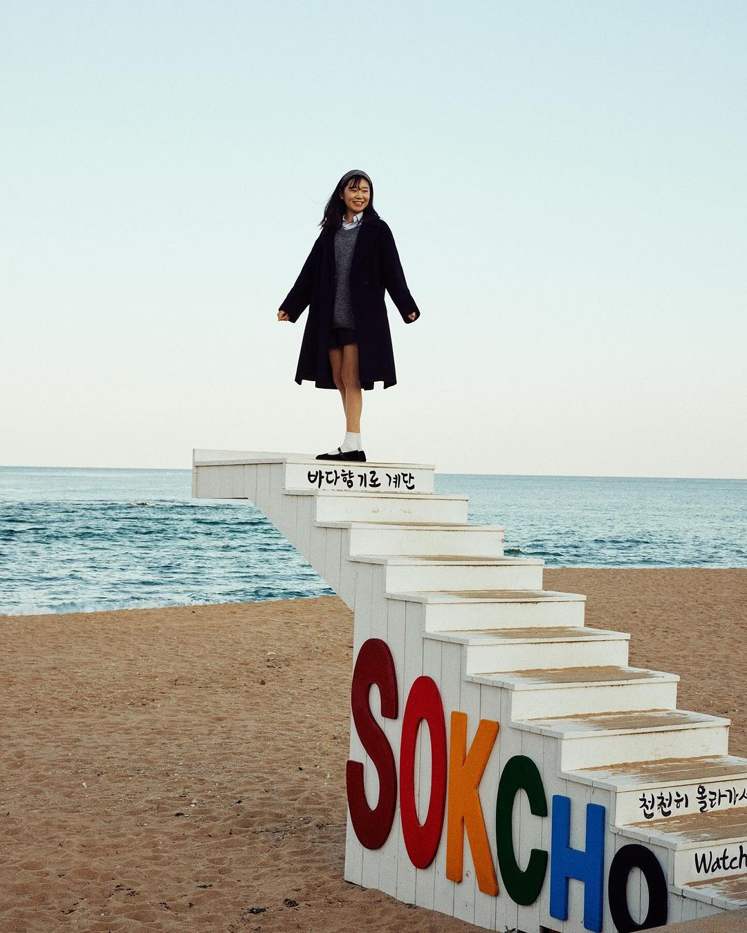 Woman standing atop a white outdoor staircase lettered SOKCHO on a sandy beach against the ocean.