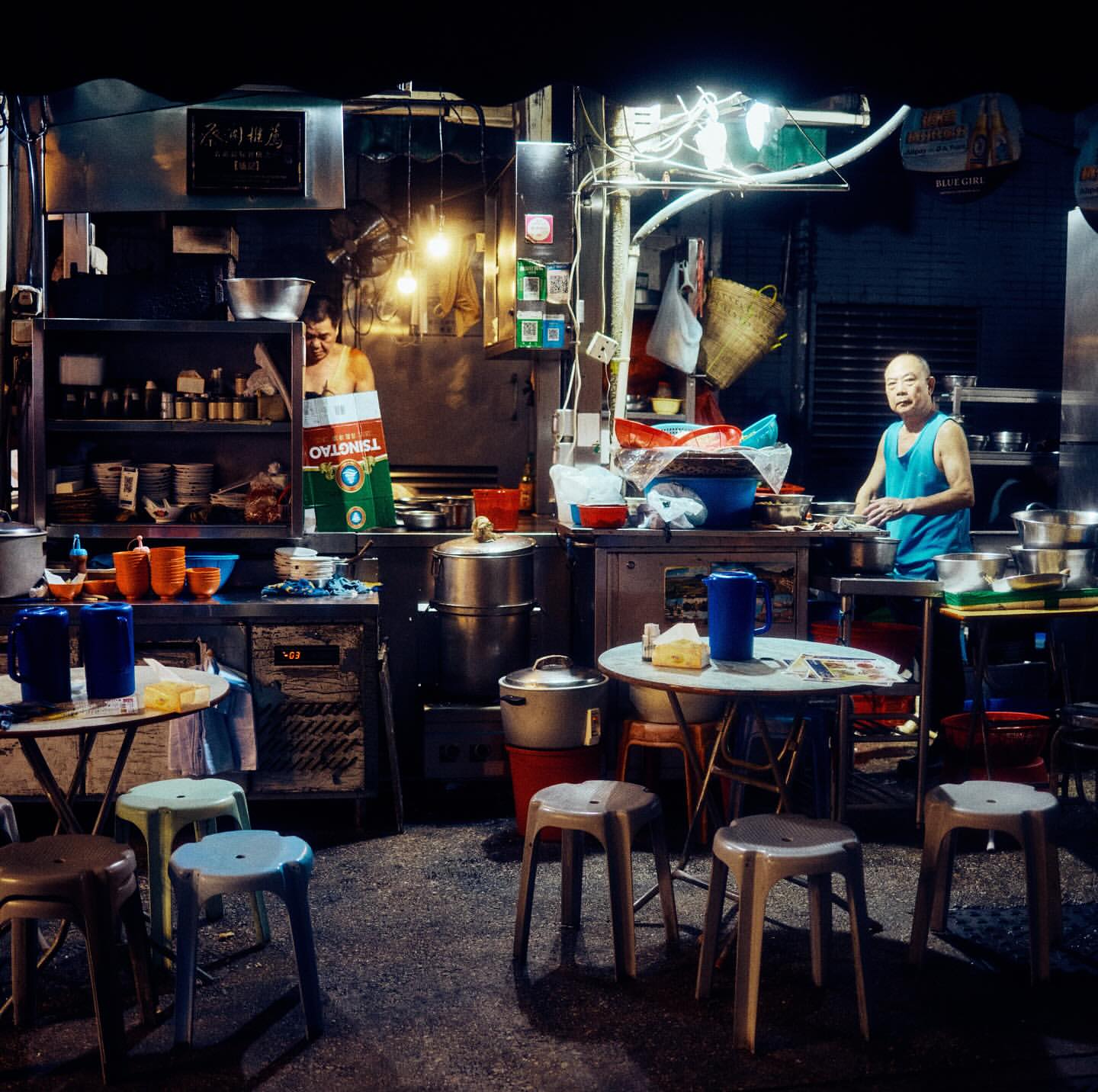 Two people prepare food at a street food stall at night, with stools and tables filling the foreground in an urban setting.
