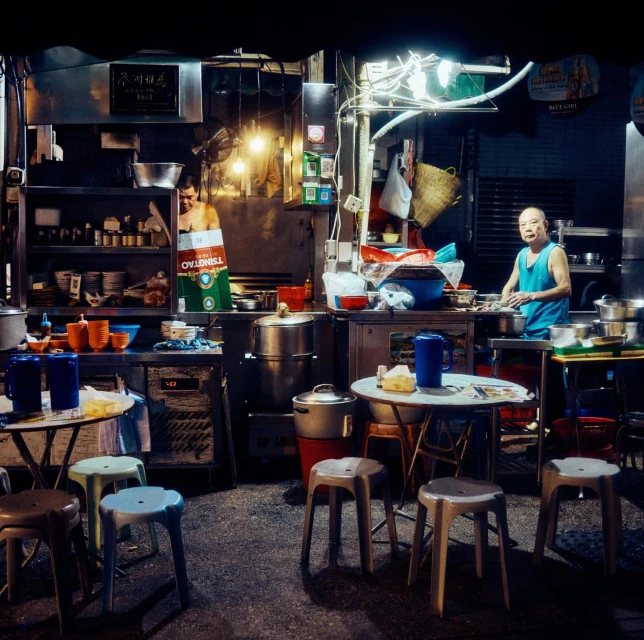 Two people prepare food at a street food stall at night, with stools and tables filling the foreground in an urban setting.